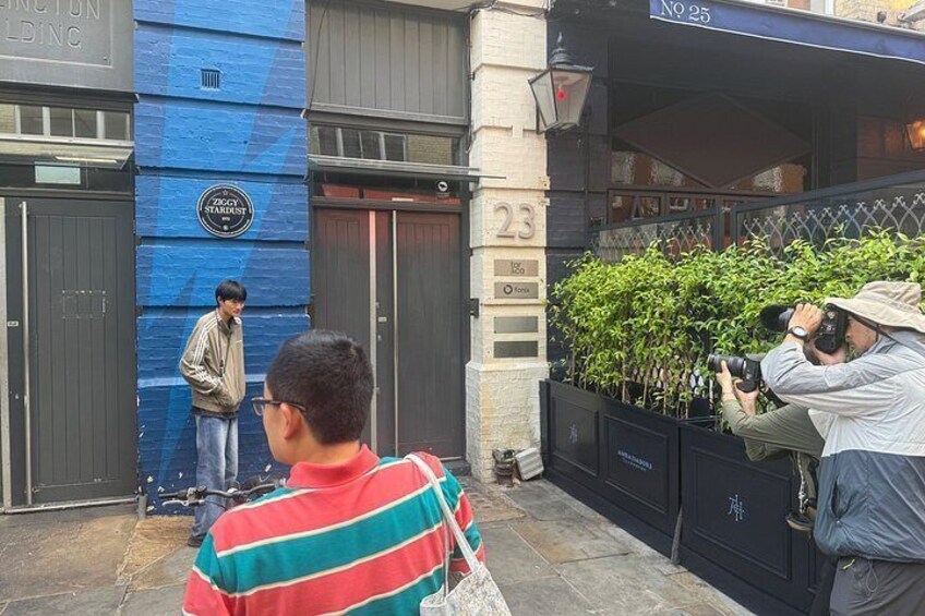 A street with a blue building, a person taking a photo, and another person standing by a gate. 2