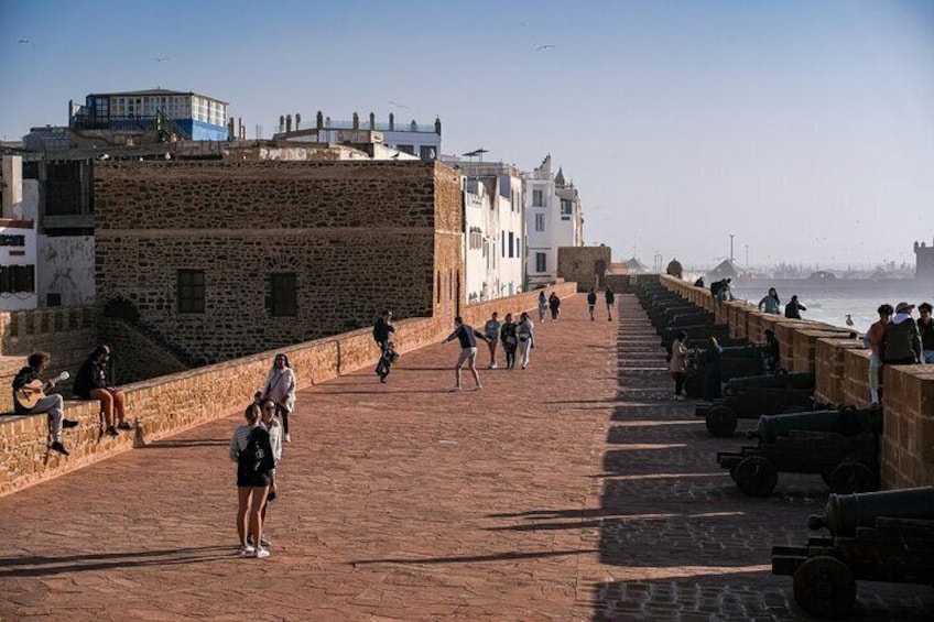 A stone-paved promenade with people walking, a row of cannons, and buildings in the background. 1