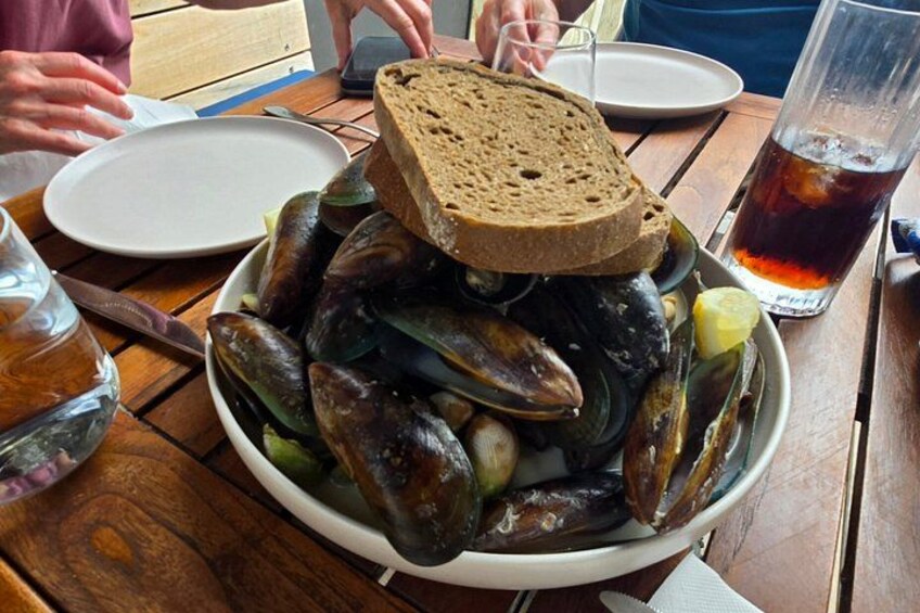 A bowl of mussels with a slice of bread on top, a glass of dark beverage, and a white plate on a wooden table. 2