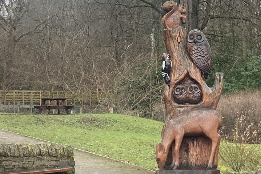 A wooden sculpture with owls and a bird perched on it, set against a backdrop of trees and a picnic table. 5