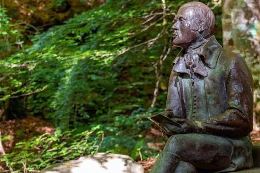 A bronze statue of a seated man with a book, surrounded by lush greenery. 3