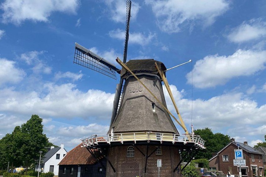 A traditional windmill with a wooden structure and a metal windlass, surrounded by buildings and trees. 4