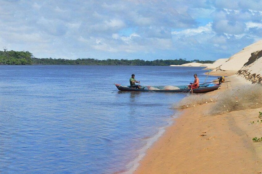 Two people in a boat on a calm lake with a sandy shore and a forest in the background. 3