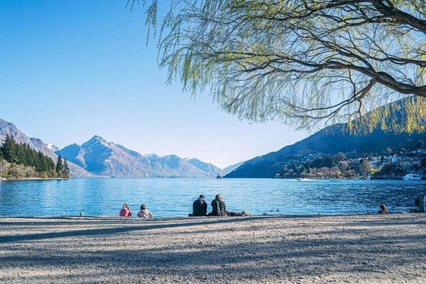 En sjö med berg i bakgrunden, en grupp människor som sitter vid stranden och ett träd med hängande grenar. 3