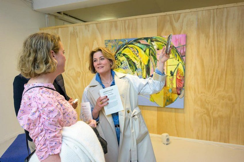 A person in a white coat holding a document and a phone, standing in front of a colorful painting. 2