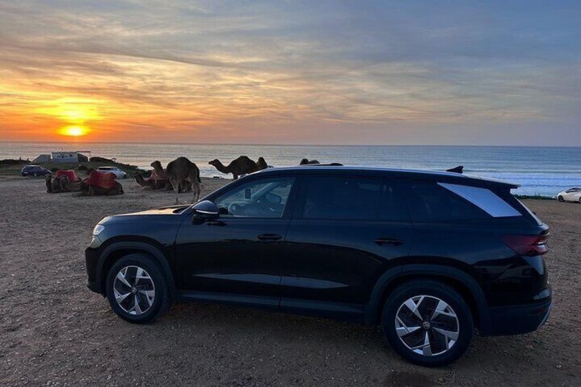 Un SUV negro estacionado en una playa al atardecer, con camellos y tiendas de campaña al fondo. 2