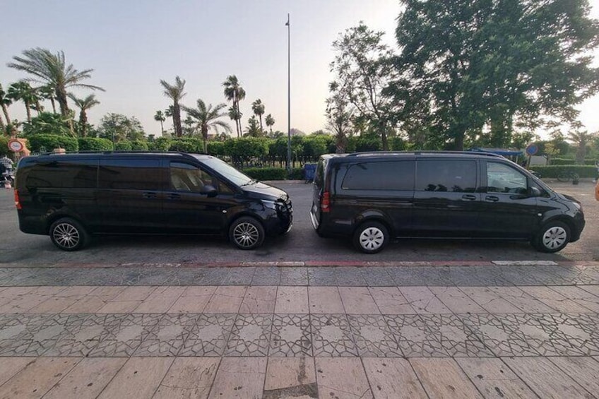 Dos furgonetas negras estacionadas sobre un pavimento de baldosas, con palmeras y un asta de banderas al fondo. 3