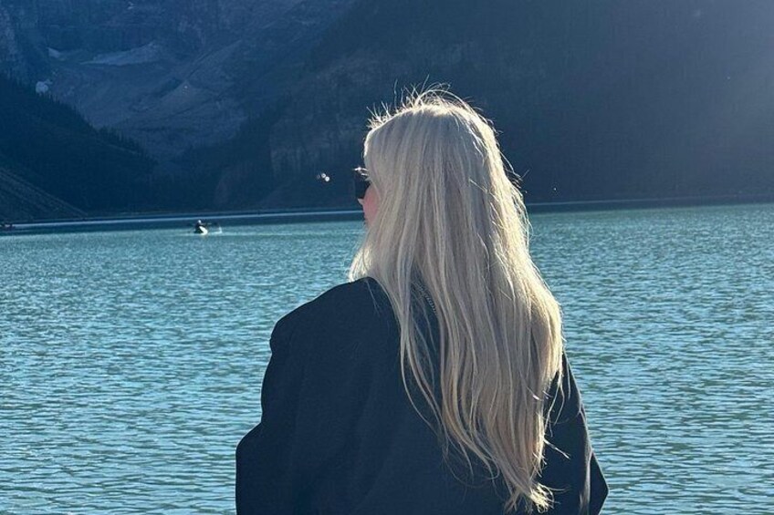 A person with long blonde hair standing by a calm lake with mountains in the background. 4