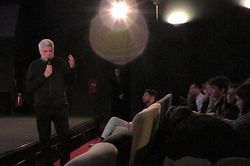 A person speaking on stage in a dimly lit auditorium with an audience seated in rows. 4