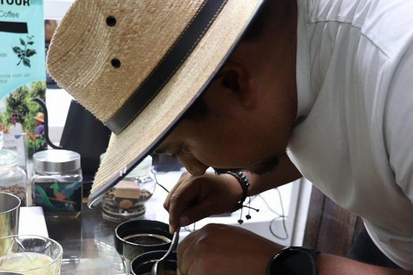 Una persona con un cappello di paglia e una camicia bianca, che tiene un cucchiaino sopra una tazza, probabilmente assaggiando del caffè. 1