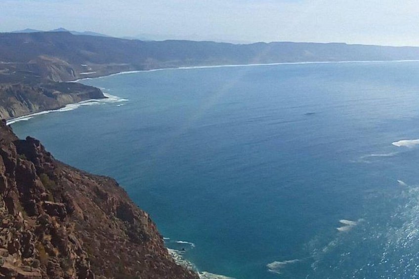 Paisaje costero con un acantilado escarpado, una vasta extensión de océano y montañas distantes bajo un cielo despejado. 3