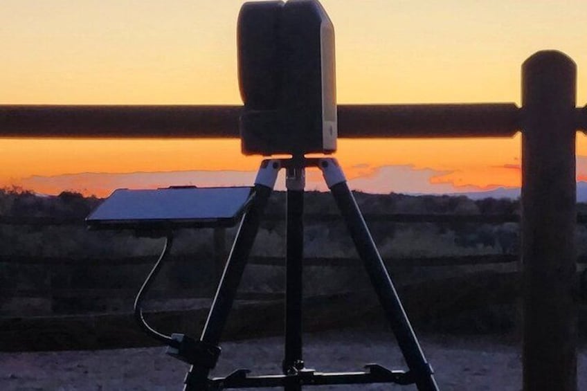 Un trípode con una cámara sobre arena, una valla de madera y un atardecer al fondo. 1