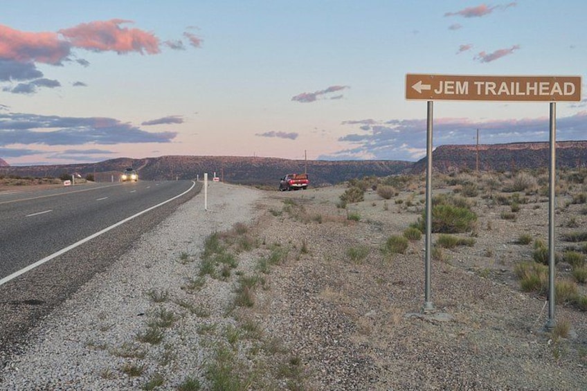 A road with a sign for JEM TRAILHEAD, a gravel area with sparse vegetation, and a distant vehicle. 5