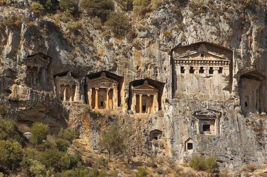 Ancient rock-cut tombs with columns and a large central structure on a hillside. 2