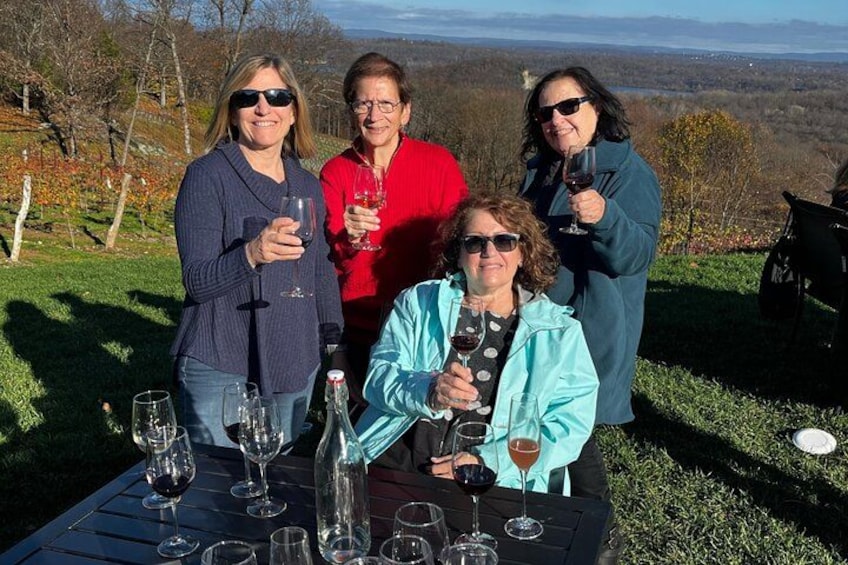 Four people standing at a table with wine glasses, outdoors with a view of trees and hills. 1
