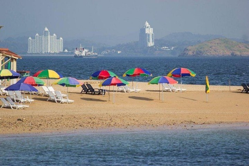 A beach with colorful umbrellas and lounge chairs, a calm sea, and distant buildings. 3