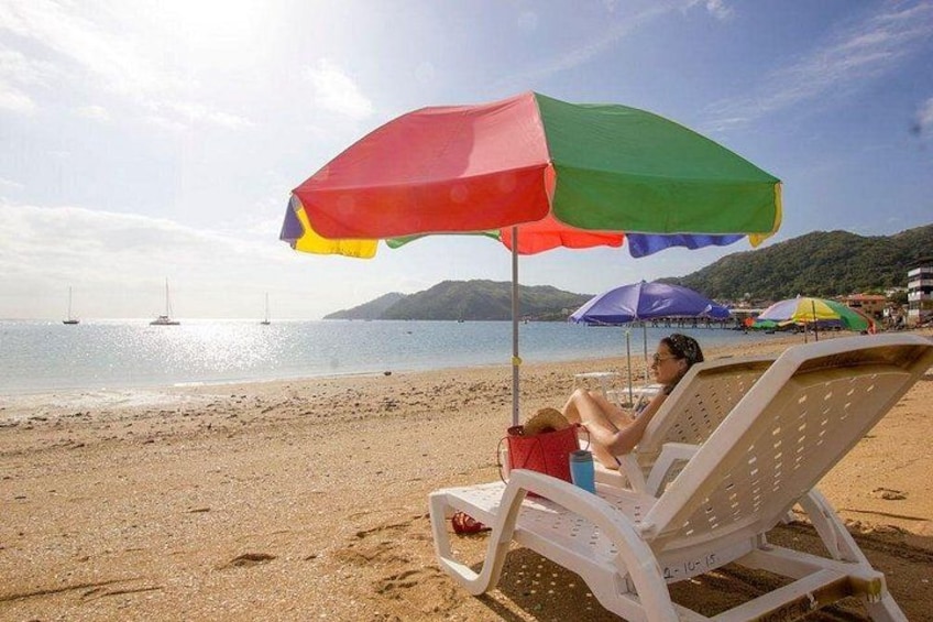 A beach with a person relaxing under a colorful umbrella, a sandy shore, and sailboats in the distance. 5