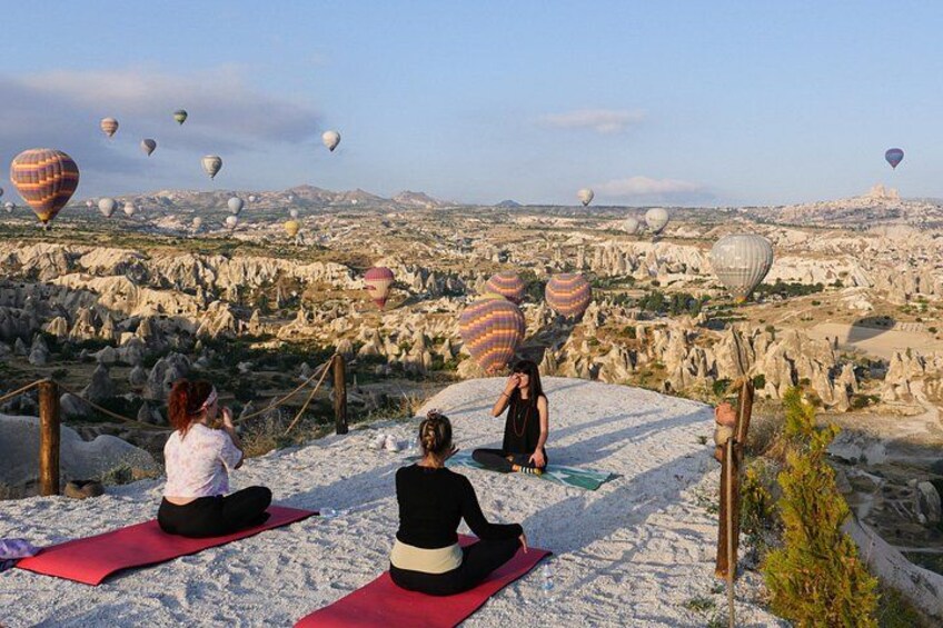 Personas meditando sobre esterillas rojas en una zona montañosa con globos aerostáticos al fondo. 1