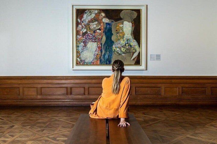 Eine Person sitzt auf einer Bank in einer Kunstgalerie und blickt auf ein großes Gemälde an der Wand. 1