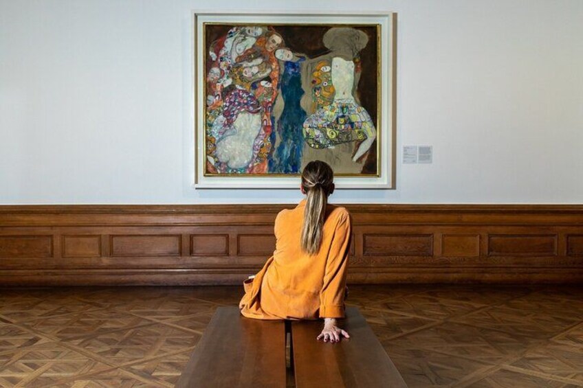A person in a yellow dress sitting on a bench in a room with a large painting on the wall. 1
