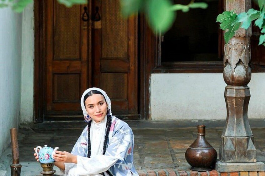 A person sitting on the ground holding a teapot, with a wooden door and a decorative vase in the background. 5