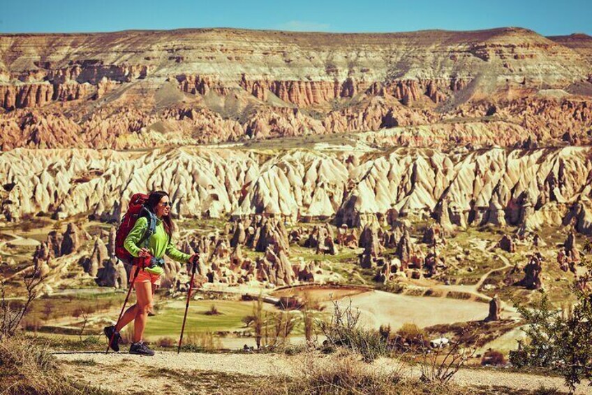 Un excursionista con chaqueta verde y pantalones cortos rosa camina sobre un sendero de tierra en un paisaje rocoso y erosionado con formaciones rocosas distintivas. 2