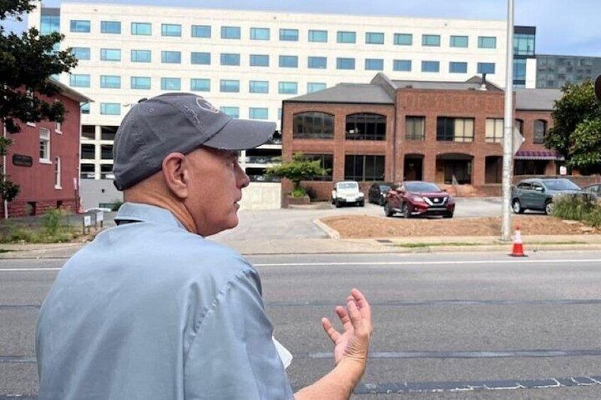 A man gesturing on a street with buildings and parked cars in the background. 3