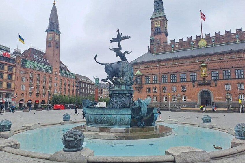 A historic building with a clock tower, a fountain with sculptures, and a flag. 3