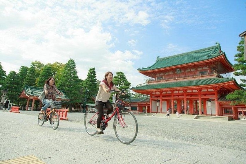 To personer cykler foran en traditionel japansk bygning med et grønt tag. 2