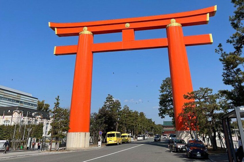 En stor, rød torii-port med to søjler og en vandret bjælke, omgivet af en klar blå himmel og bybygninger. 3