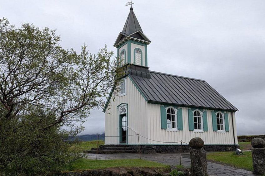 A white church with green trim and a steeple, surrounded by trees and a stone wall. 4