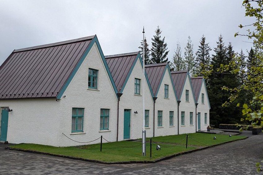 A row of white buildings with green trim and brown roofs, surrounded by a paved area and trees. 5