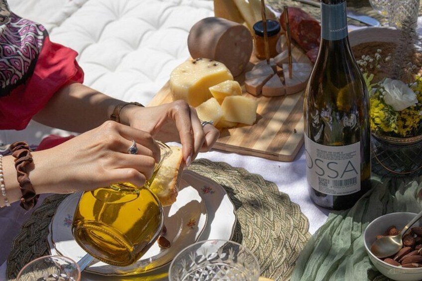 Una persona versa olio d'oliva sul pane, circondato da formaggi, salumi e una bottiglia di vino su un tavolo da picnic. 5