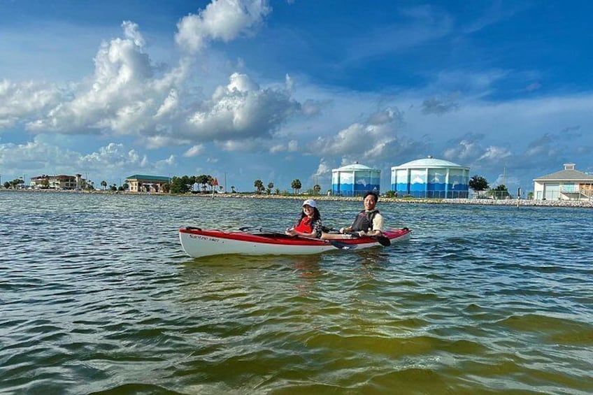 Deux personnes dans un canoë rouge et blanc sur une étendue d’eau calme, avec des bâtiments et une structure en forme de dôme en arrière-plan. 1