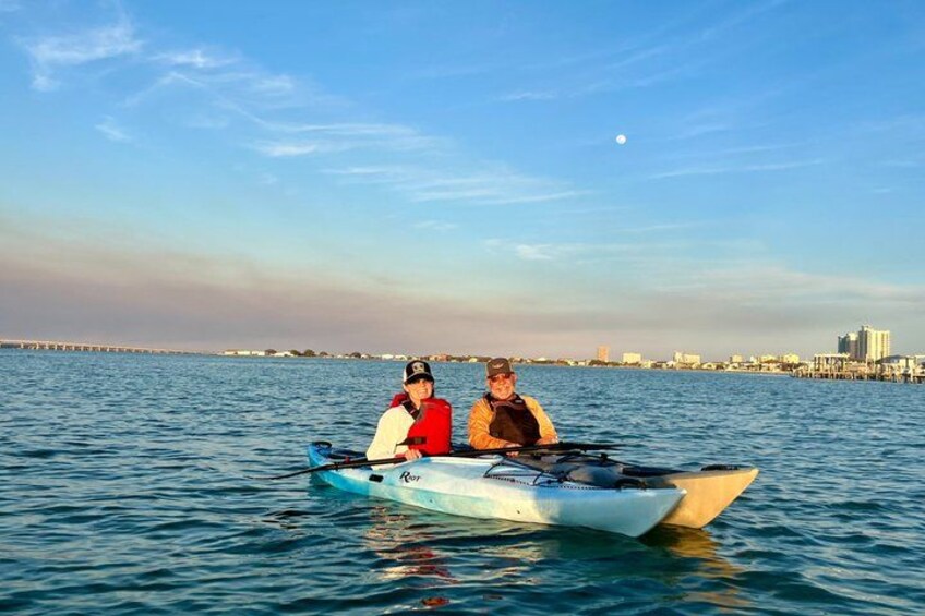 Two people in a small boat on calm water with a city skyline in the background. 1