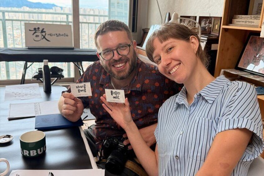 Two people holding cards with Korean text, a coffee cup, and a camera on a desk. 1