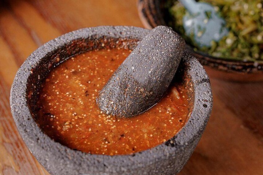 A stone mortar with a pestle, a bowl of red sauce, and a bowl of green sauce in the background. 3