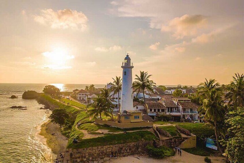 Un pueblo costero con un faro destacado, palmeras y casas junto al mar al atardecer. 1