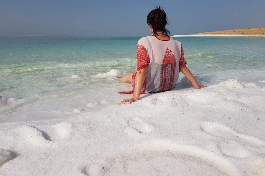 A person sitting on a sandy beach with clear turquoise water and a distant shoreline. 2