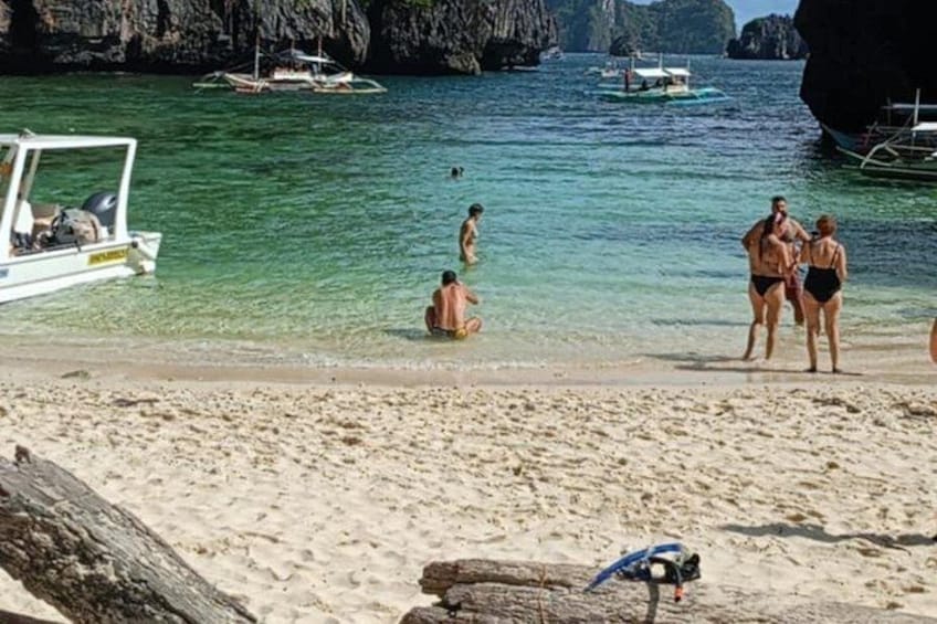 A beach with people sunbathing and swimming, boats in the water, and a rocky coastline. 3