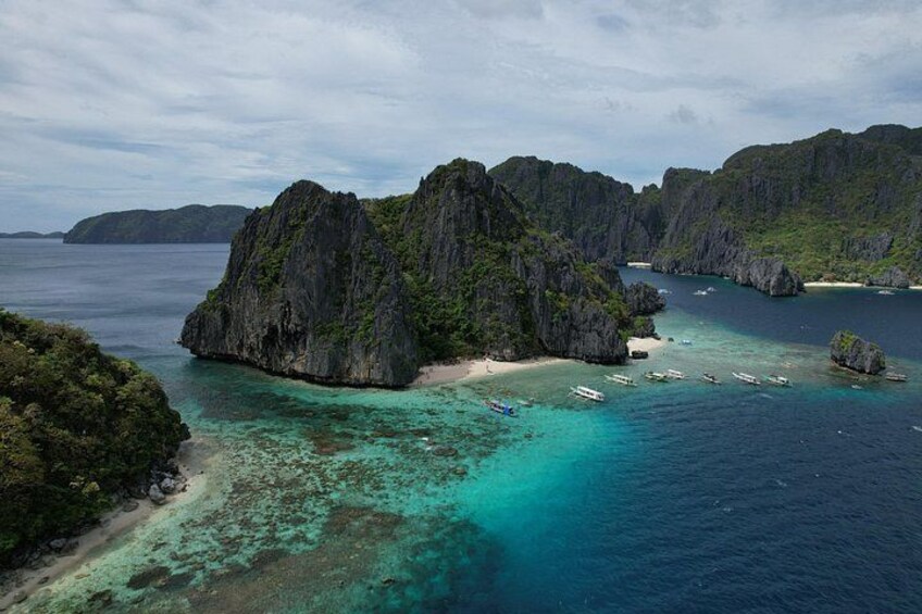 A coastal landscape with a prominent rocky island, clear turquoise waters, and several boats anchored near the shore. 1
