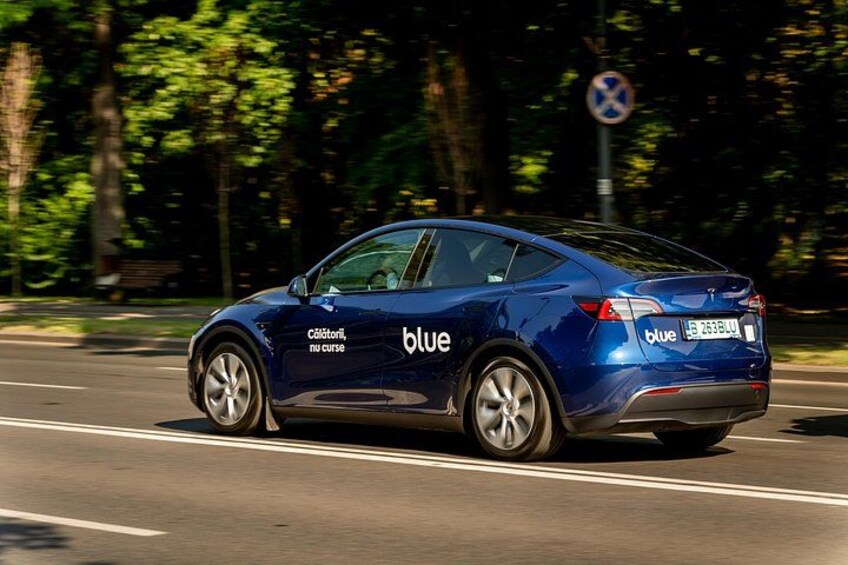 A blue car with 'blue' and 'Coloriati, nu curve' on the side, driving on a road with trees and a speed limit sign in the background. 3