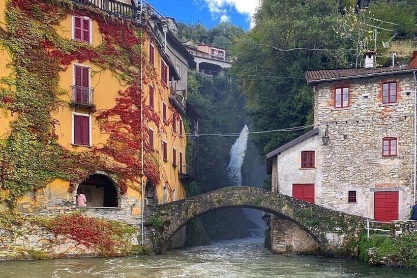 A stone bridge over a river, buildings with red shutters, and a waterfall in the background. 3