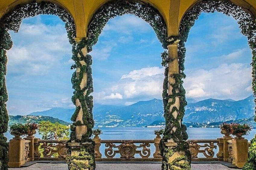 A balcony with ornate columns and greenery, overlooking a lake and mountains. 4