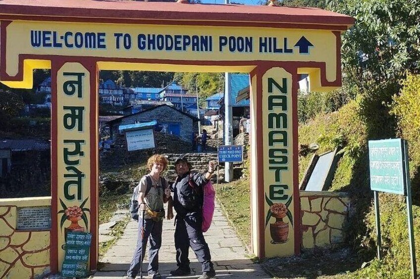 A welcome sign at Ghodepanji Poon Hill with a directional arrow, a couple taking a photo, and a signboard with text in a local language. 3