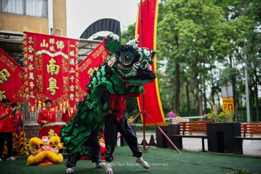 A lion dance performance with performers in green and red costumes, banners with Chinese characters, and a stage set outdoors. 3