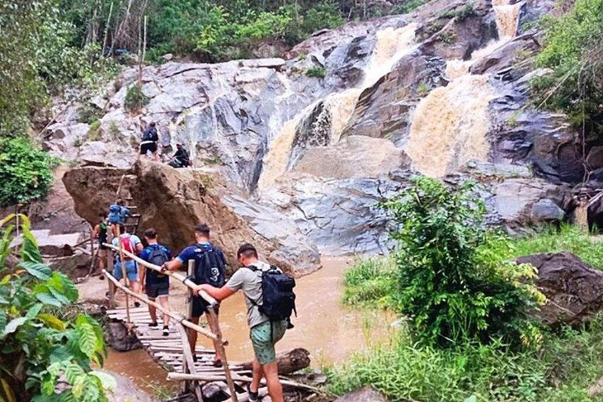 A group of hikers crossing a wooden bridge over a river in a forested area with a waterfall in the background. 1