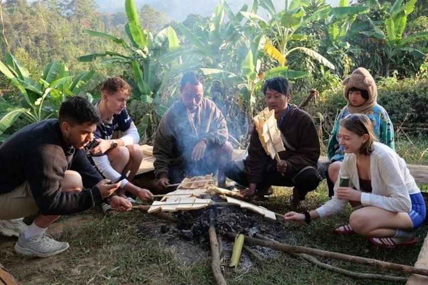 A group of people around a campfire in a natural setting with greenery in the background. 4