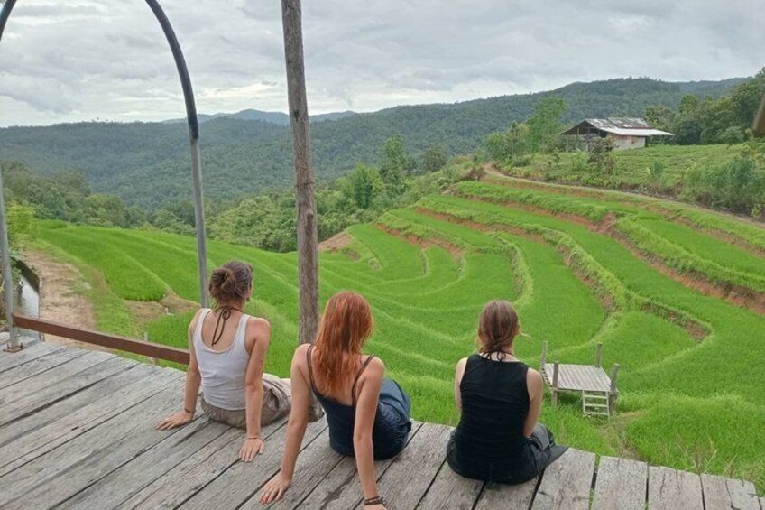 Three people sitting on a wooden deck overlooking terraced rice fields and a house in the distance. 4