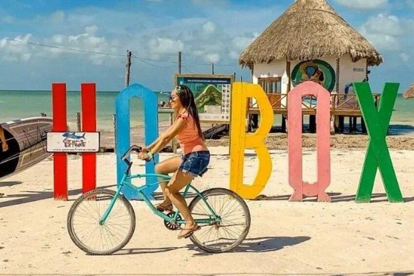 Une personne à vélo sur une plage de sable, avec de grandes lettres colorées formant le mot « BOX » en arrière-plan. 1